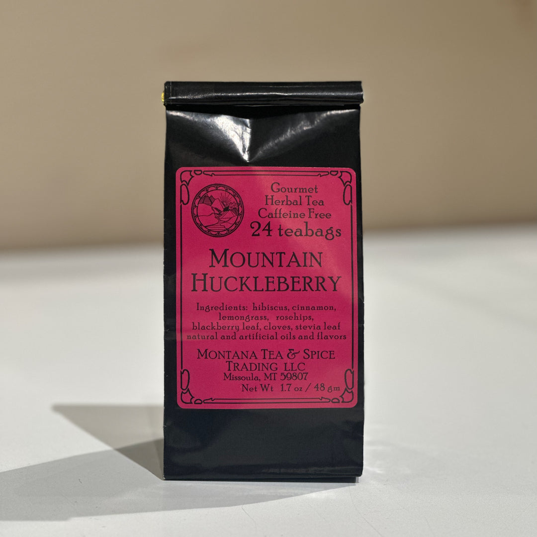 A black stand-up pouch of Mountain Huckleberry herbal tea by Montana Tea &amp; Spice Trading LLC, featuring a pink label, set against a neutral beige background.