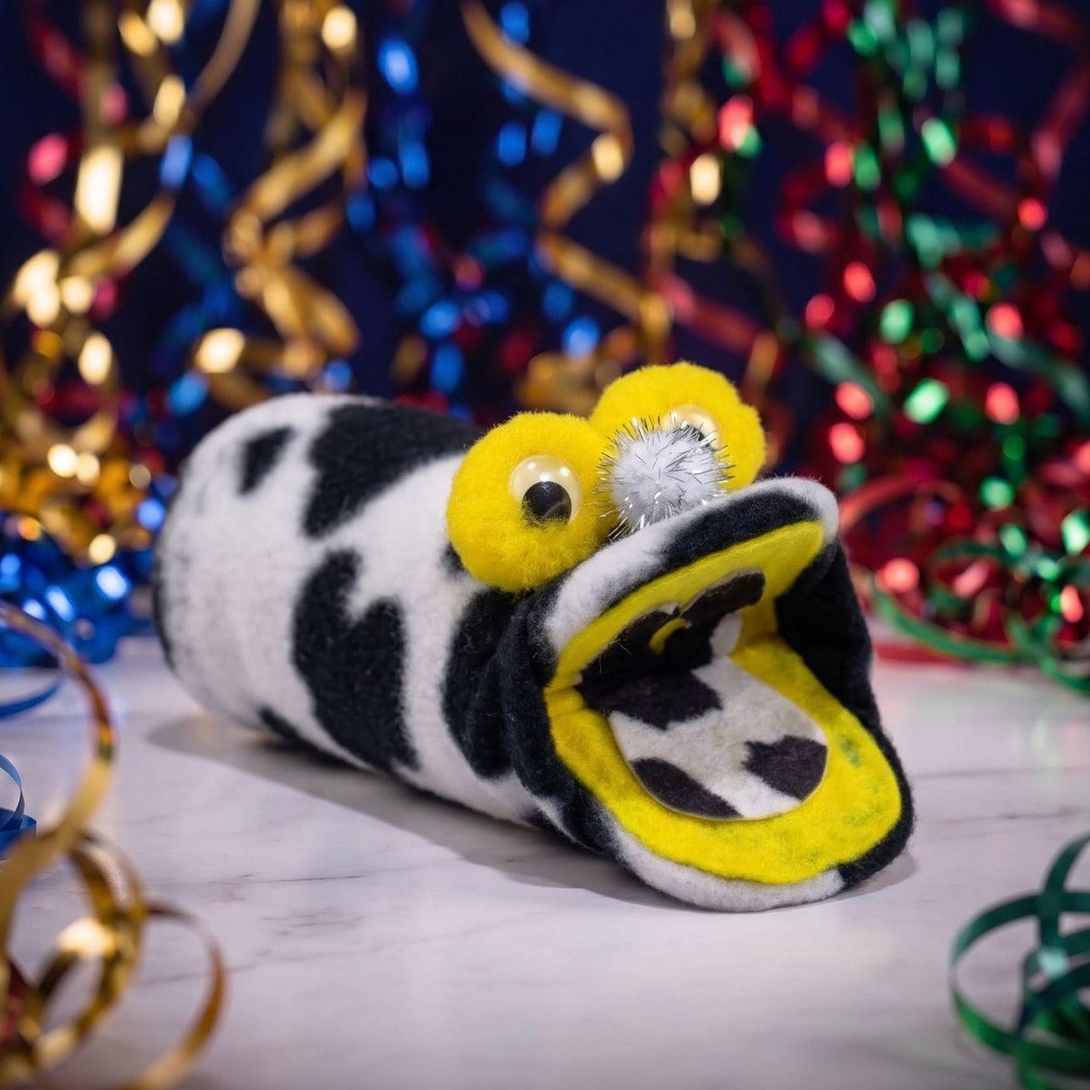 A plush novelty pillow shaped like a cow, with its mouth wide open to reveal a small plush snake inside. The pillow features a black and white cow print with bright yellow accents on the eyes and mouth interior, set against a festive background of colorful tinsel.