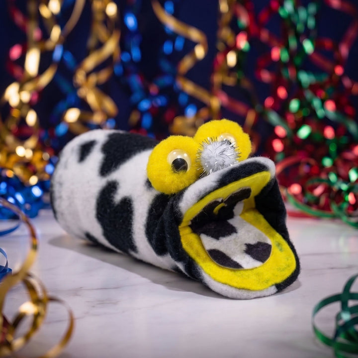 A plush novelty pillow shaped like a cow, with its mouth wide open to reveal a small plush snake inside. The pillow features a black and white cow print with bright yellow accents on the eyes and mouth interior, set against a festive background of colorful tinsel.