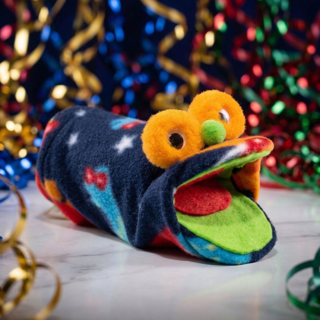 A plush handmade puppet, with orange yarn eyes, a green nose, and an open mouth showing a red tongue and green inner mouth. The toy is photographed on a white marble-like surface with festive gold and red streamers in the background.