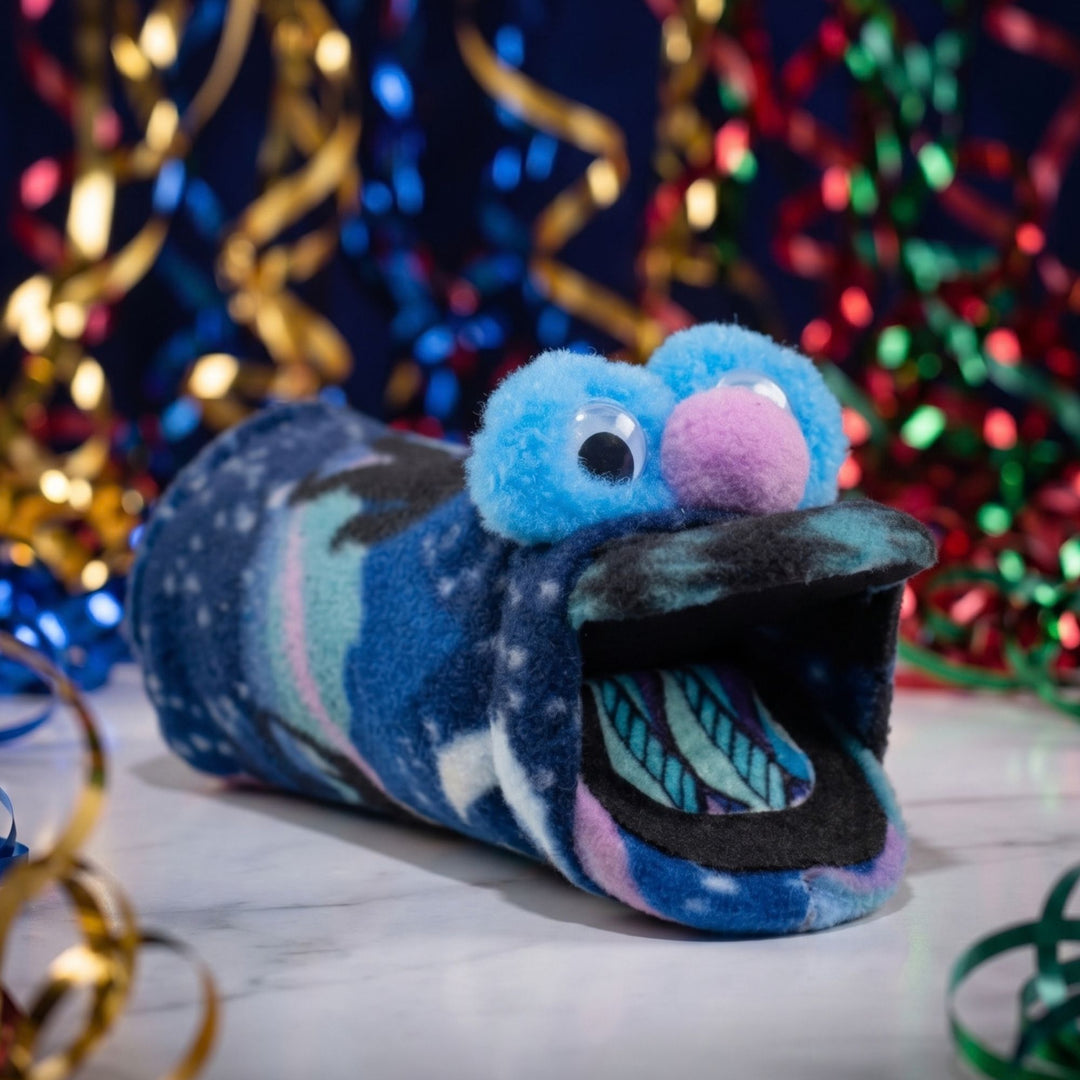 A plush hand made puppet with a blue fuzzy head, large googly eyes, a pink nose, and a galaxy-patterned body, shown on a marble surface with festive tinsel in the background.