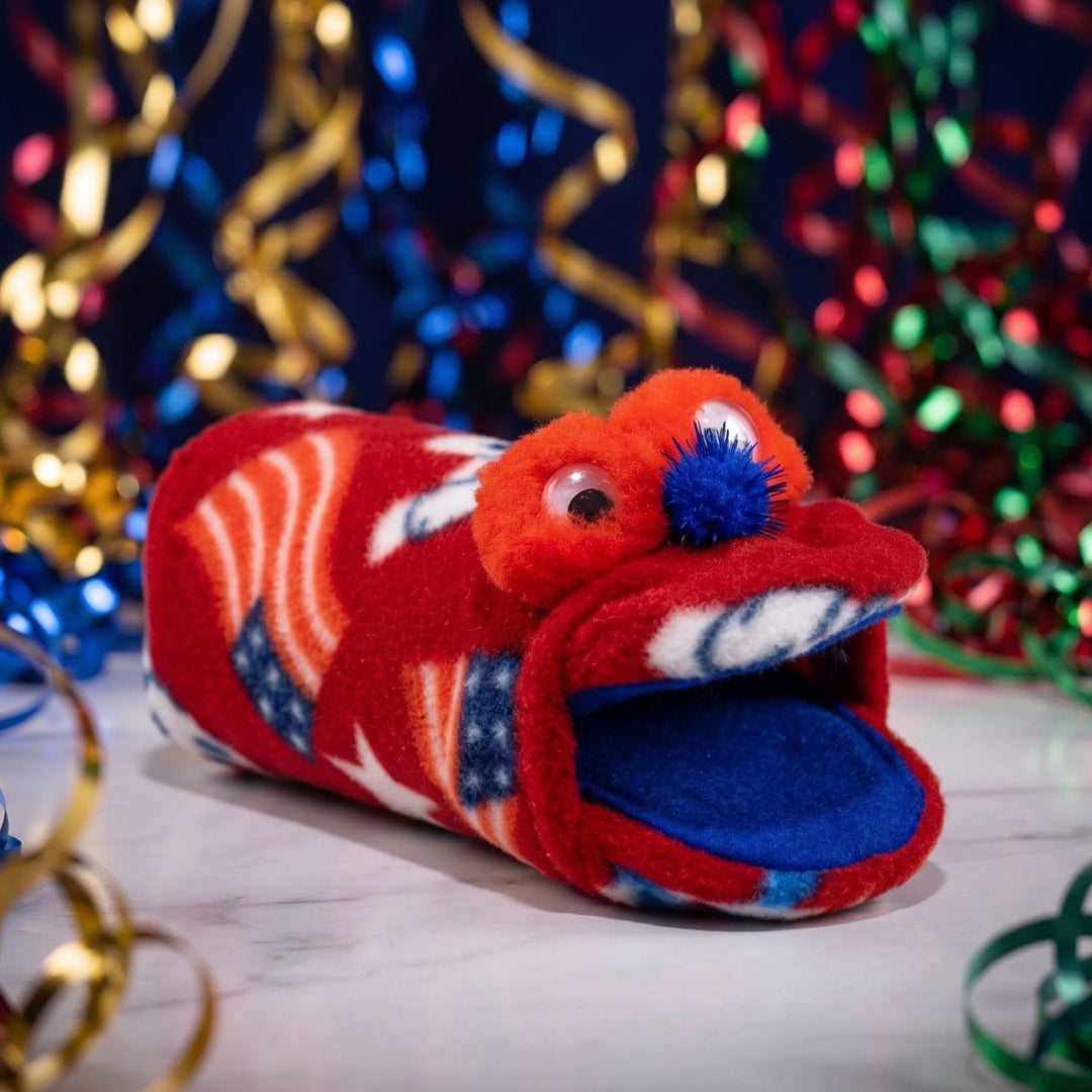 A plush handmade puppet, made from red fleece with stars and stripes pattern, featuring googly eyes, a blue pom-pom nose, and a blue inner mouth, set against a festive background with colorful tinsel.