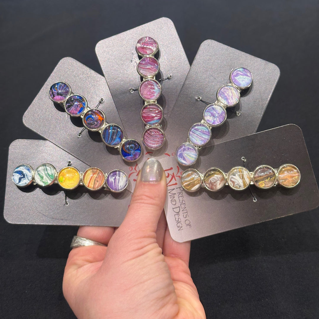 Five acrylic hand poured barrettes with swirling colors on a black background, made in Montana