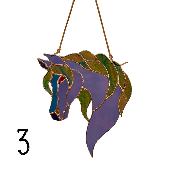 A stained glass horse head ornament with purple, green, and blue sections outlined in gold, suspended by a gold string against a white background, with a black number 3 in the lower left corner.