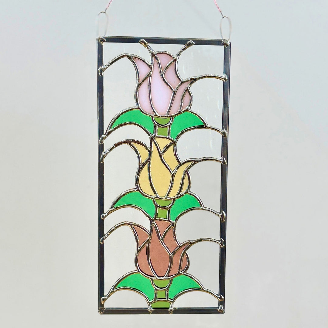A rectangular stained glass hanging panel featuring three tulip buds in pink, yellow, and peach tones with green leaves, outlined in dark metal and suspended by two small hanging loops at the top.