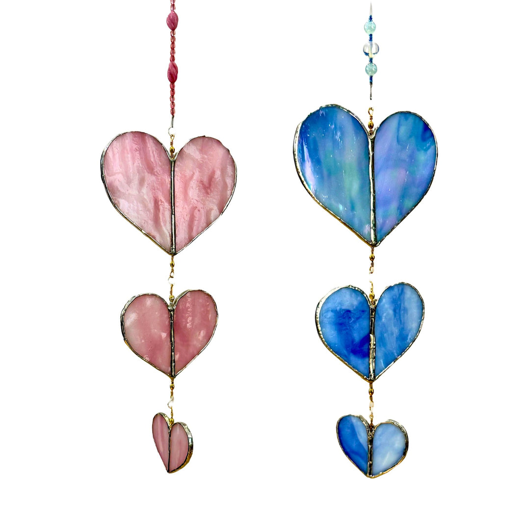 Two hanging heart-shaped stained glass suncatchers with gold metal frames on a white background. The left suncatcher has three pink marbled heart panels with a pink beaded cord, and the right suncatcher has three blue marbled heart panels with a light blue beaded cord.