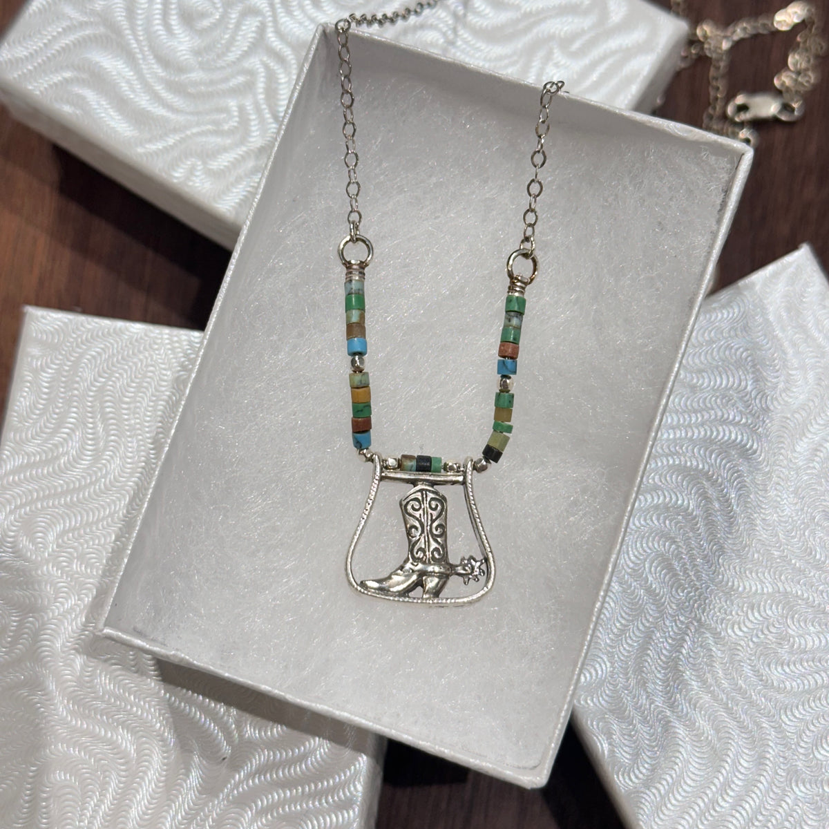 A sterling silver boot pendant and turquoise necklace in a white jewelry box on a dark wood background.