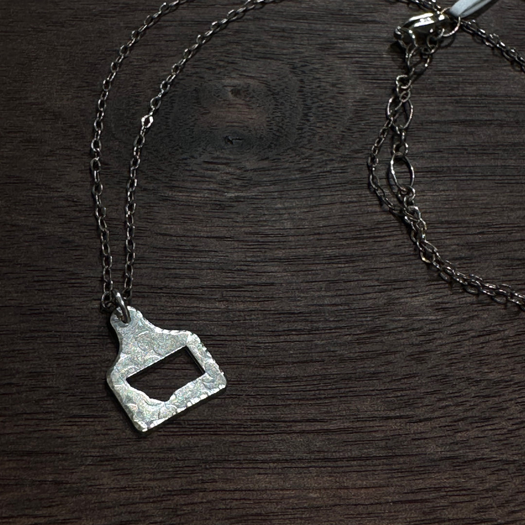 A silver cow tag pendant necklace with a hammered, textured finish and a Montana shape cutout, shown on a dark wood background.