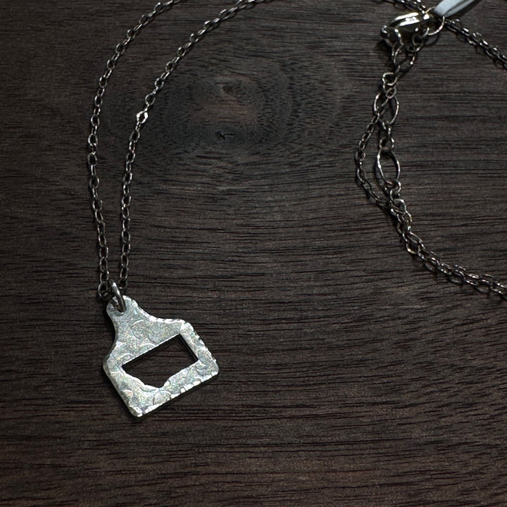 A silver cow tag pendant necklace with a hammered, textured finish and a Montana shape cutout, shown on a dark wood background.