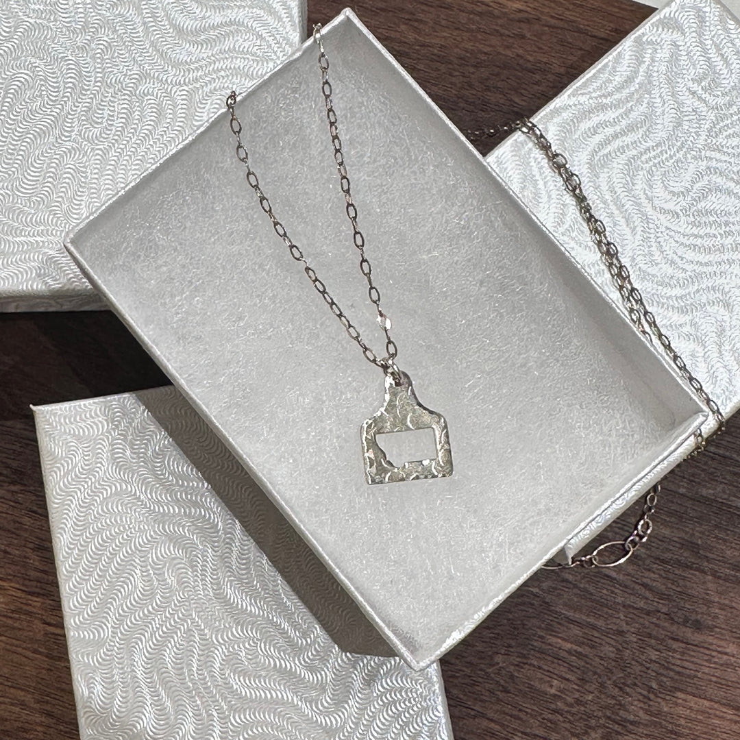 A silver cow tag pendant necklace with a hammered, textured finish and a Montana shape cutout, shown in a white jewelry box on a dark wood background.