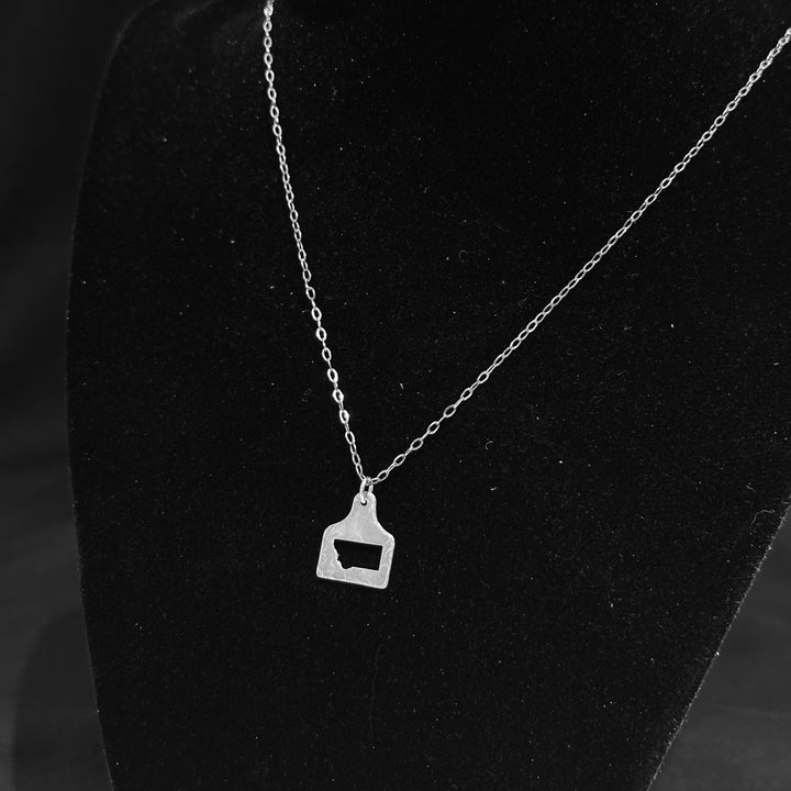 A silver cow tag pendant necklace with a hammered, textured finish and a Montana shape cutout, shown on a black background