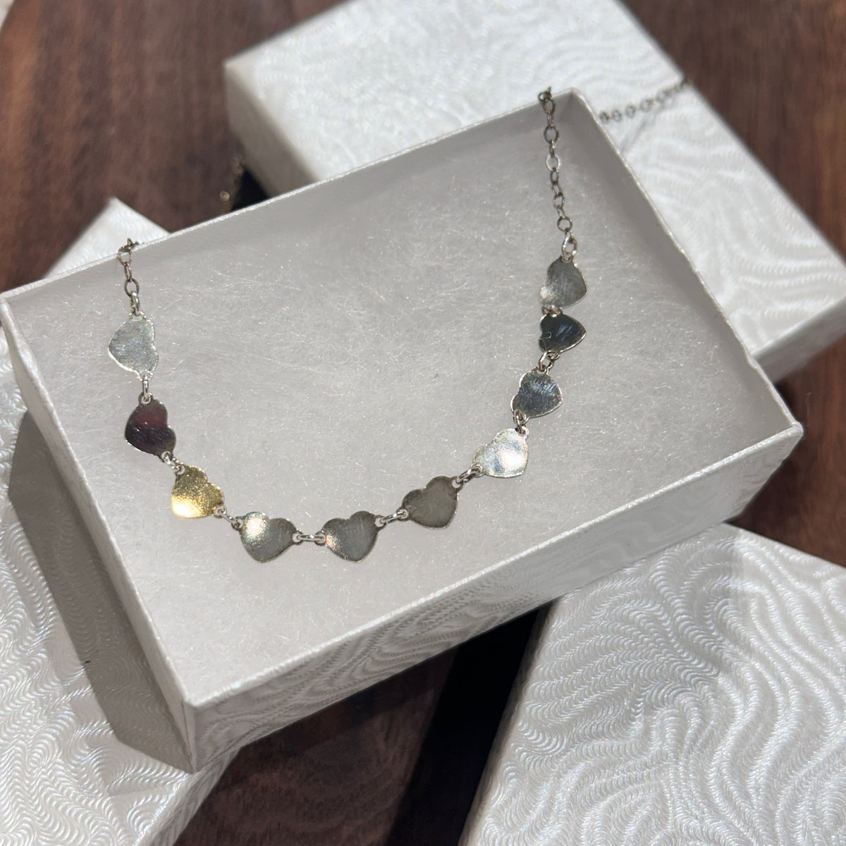 A strand of sterling silver hearts on a silver chain necklace displayed in a white jewelry box on a dark wood background