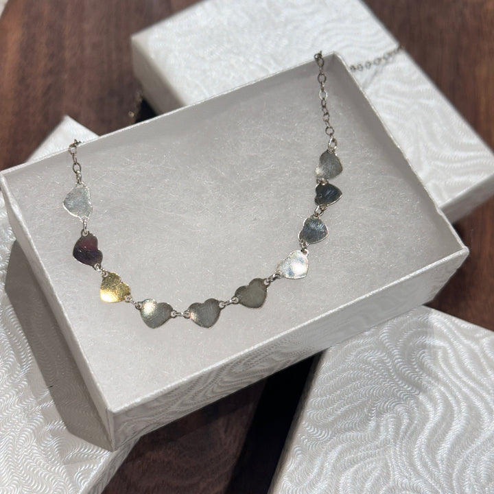 A strand of sterling silver hearts on a silver chain necklace displayed in a white jewelry box on a dark wood background