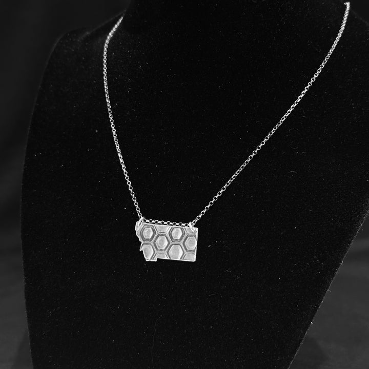 A sterling silver pendant in the shape of Montana with honeycomb engraving on a silver chain. Displayed on a black background