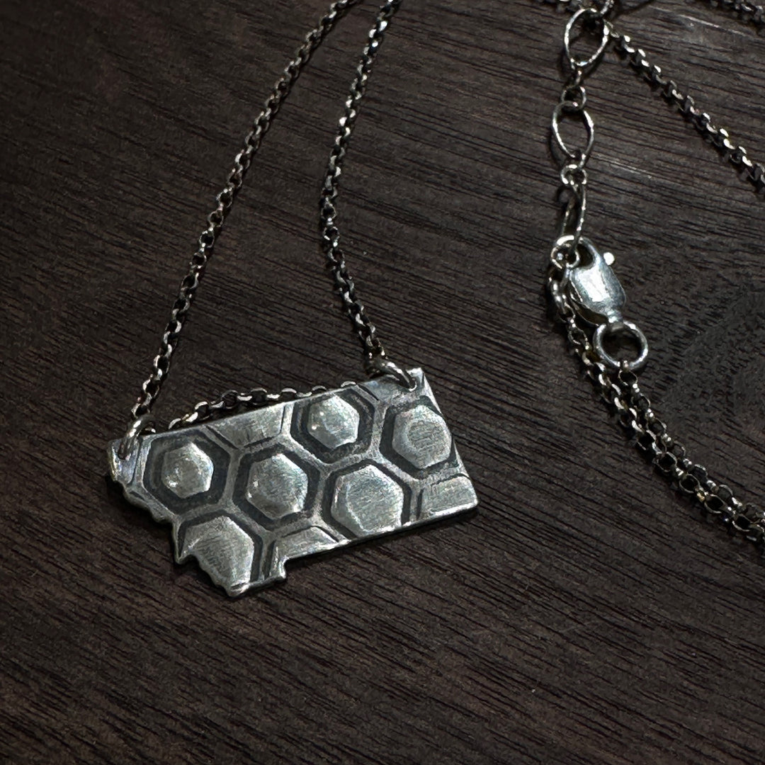 A sterling silver pendant in the shape of Montana with honeycomb engraving on a silver chain. Displayed on a dark wood background