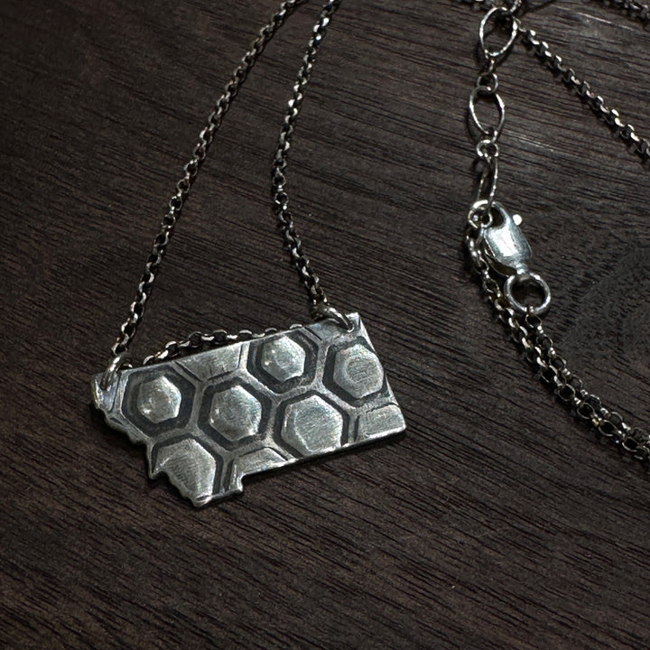 A sterling silver pendant in the shape of Montana with honeycomb engraving on a silver chain. Displayed on a dark wood background
