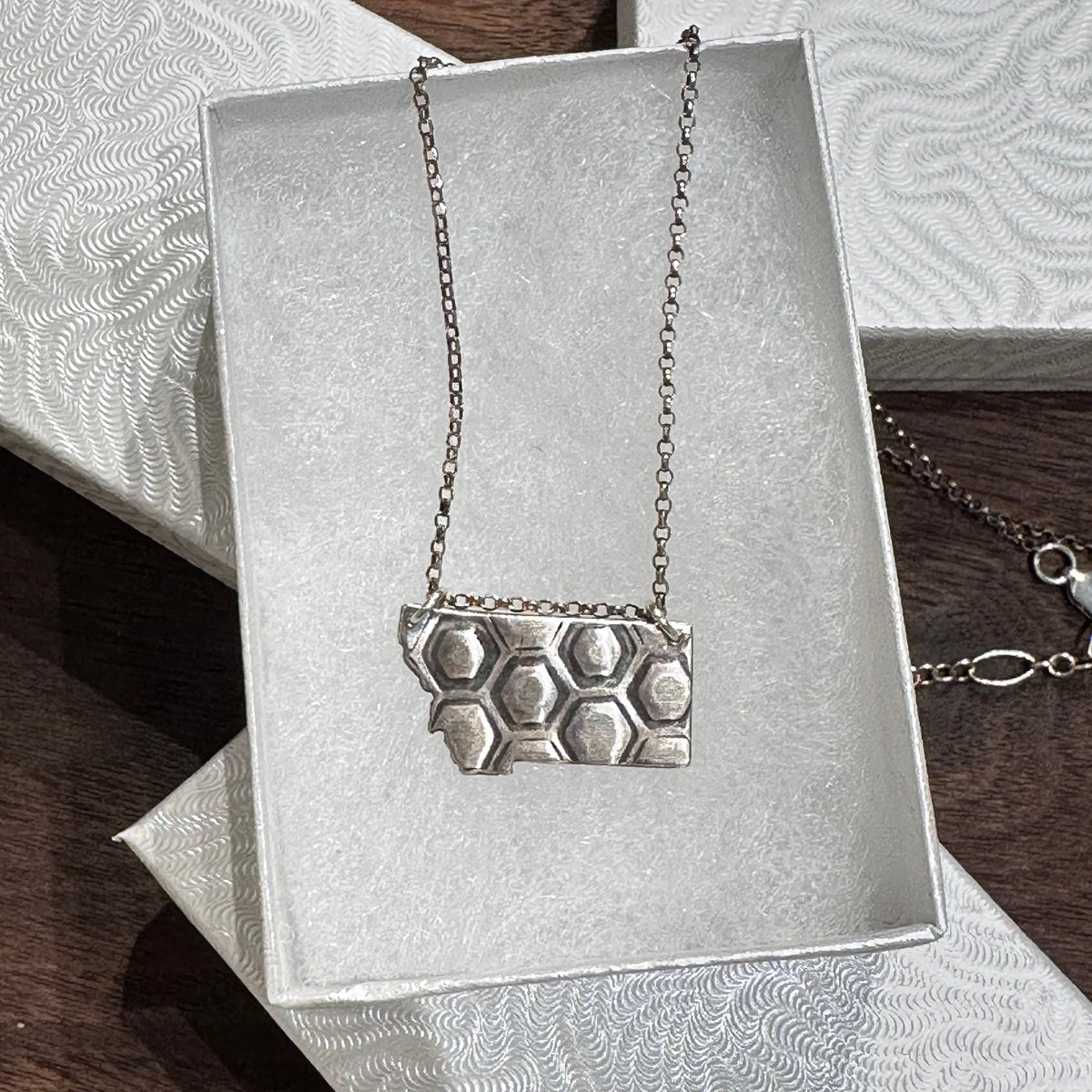 A sterling silver pendant in the shape of Montana with honeycomb engraving on a silver chain. Displayed in a white jewelry box on a dark wood background