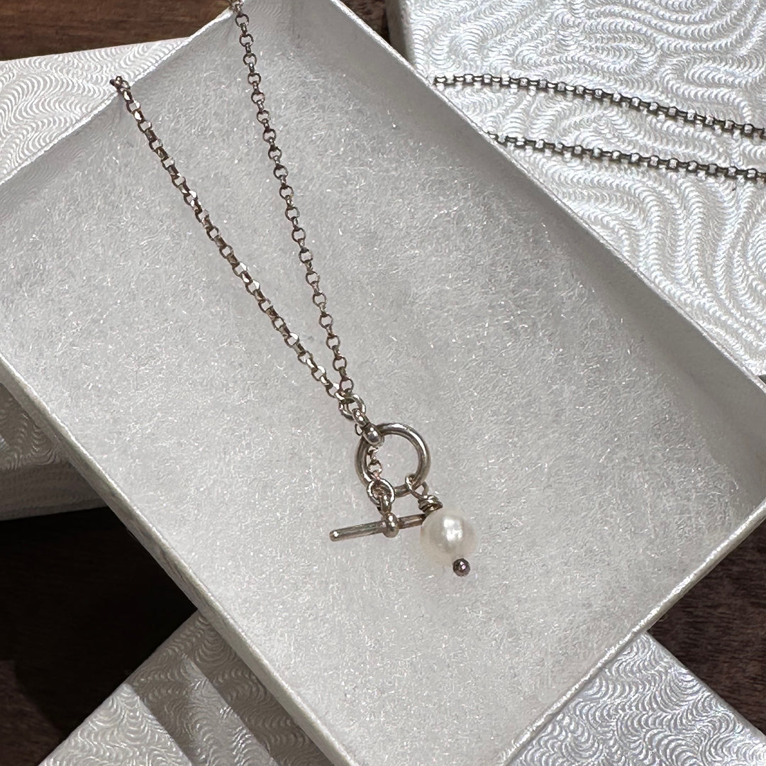 A sterling silver toggle clasp necklace with a small white pearl bead, displayed inside a textured white jewelry box.
