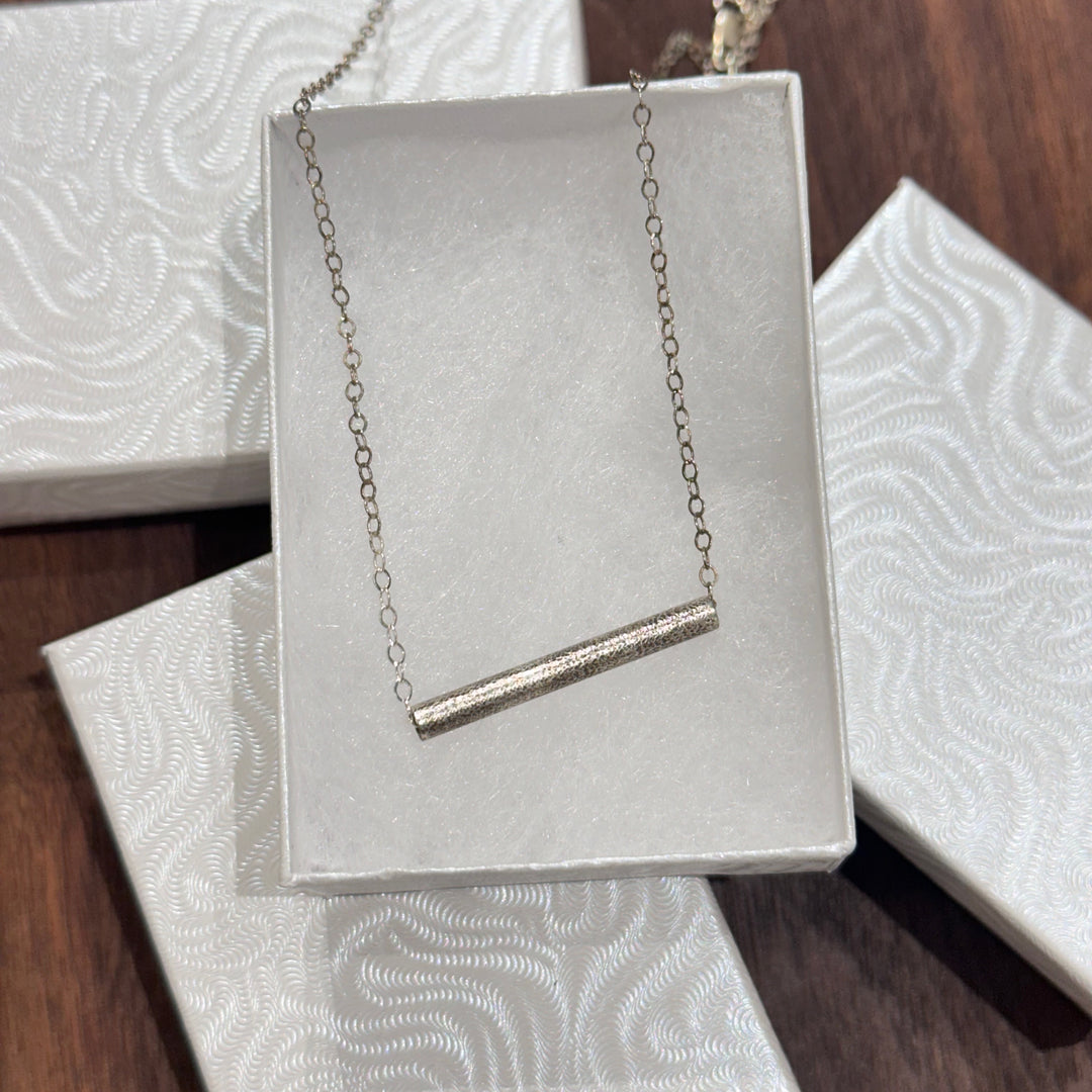 A sterling silver textured tube on a silver chain necklace displayed in a white jewelry box on a dark wood background
