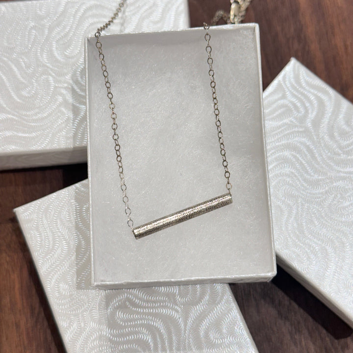 A sterling silver textured tube on a silver chain necklace displayed in a white jewelry box on a dark wood background