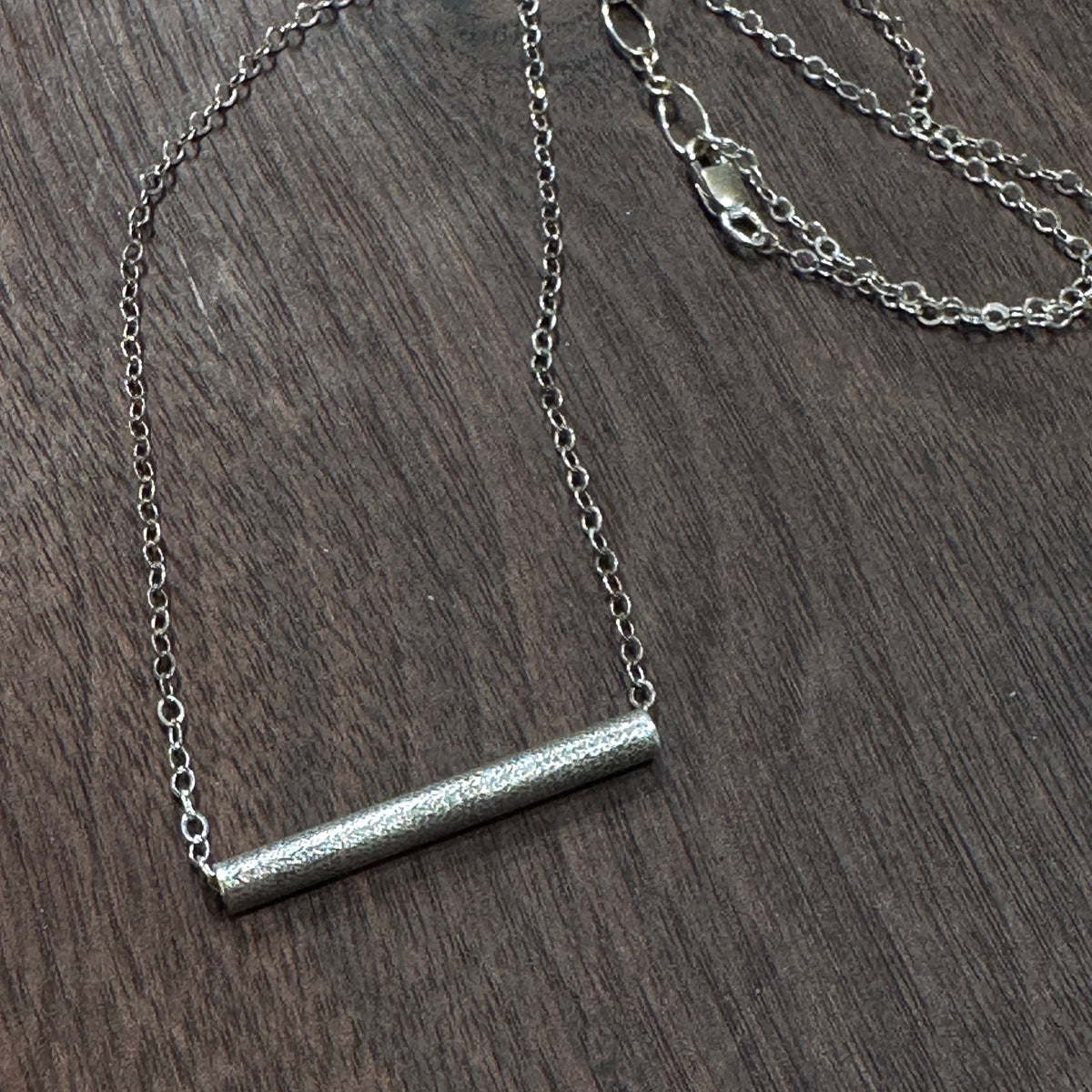 A sterling silver textured tube on a silver chain necklace on a dark wood background