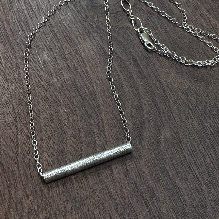 A sterling silver textured tube on a silver chain necklace on a dark wood background