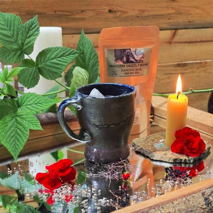 A dark ceramic mug filled with tea sits on a reflective tray beside an orange pouch of Micro blend Snoozin Grizzly tea, with lit candles, red roses, green leaves, and a rustic wood plank background.
