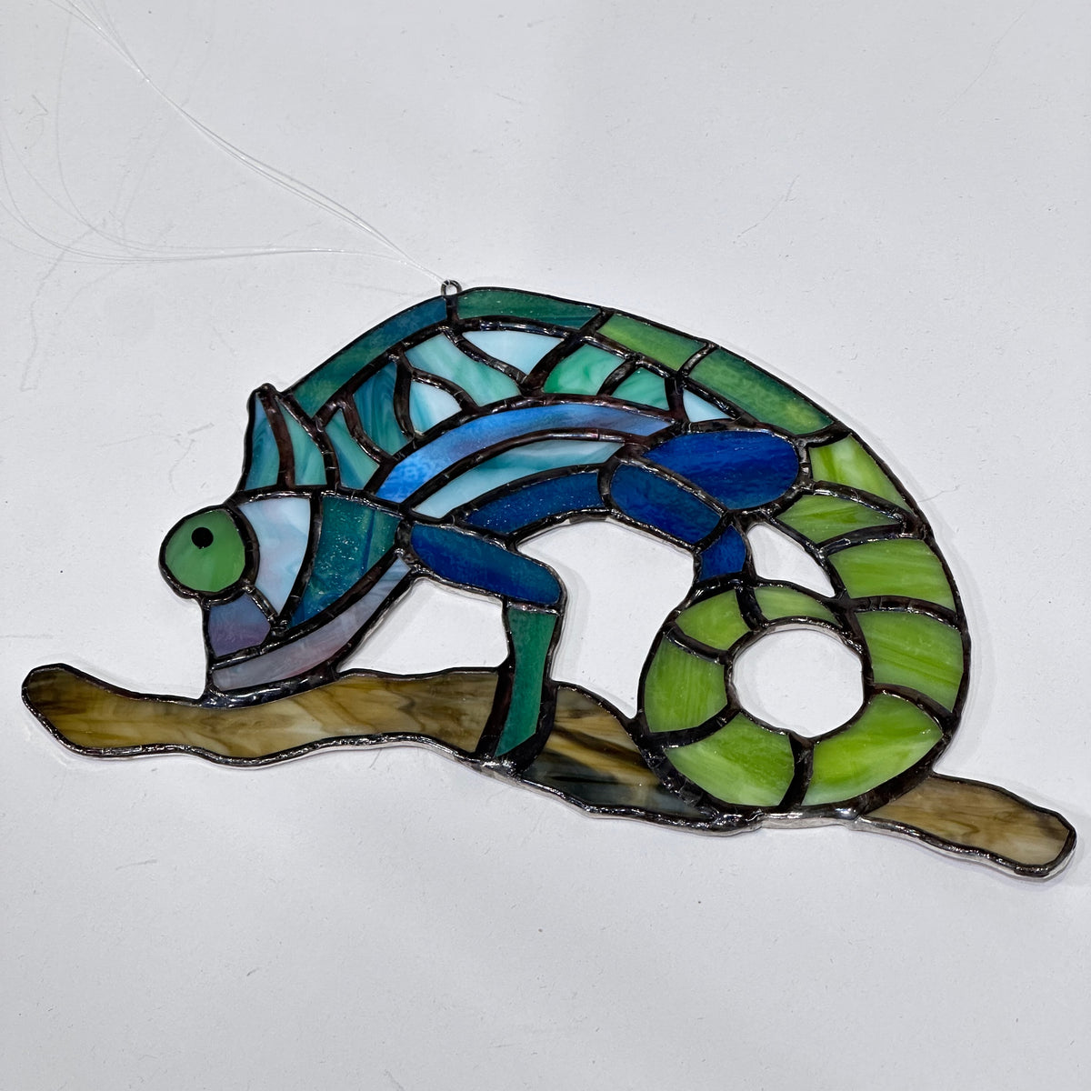 A stained glass chameleon in green, teal and blue glass on a brown glass stick, laying flat, made in montana usa