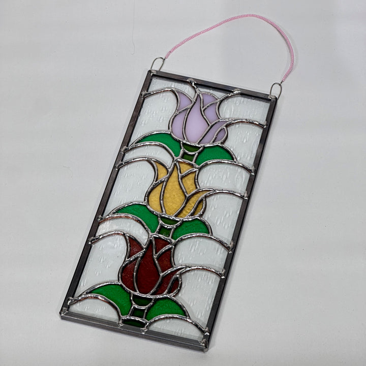 A stained glass piece with three stacked tulips in rose, yellow and pink glass in a rectangular frame.