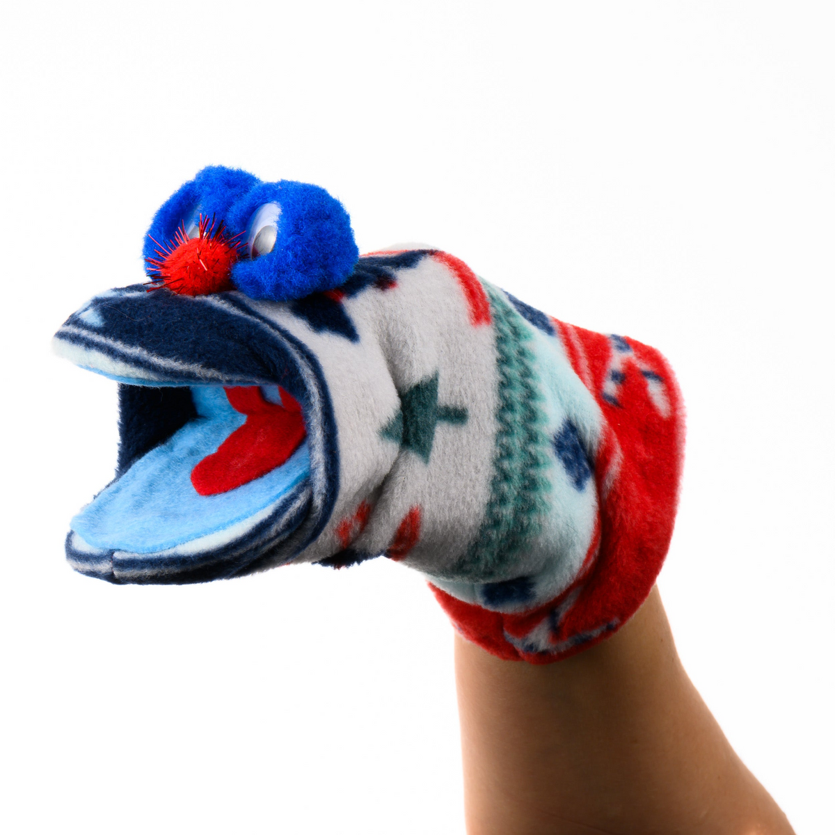 A plush sock puppet hand with a patterned white, red, and blue knit body, a blue pom-pom eye, and an open mouth showing a blue interior, worn on a human hand against a white background.