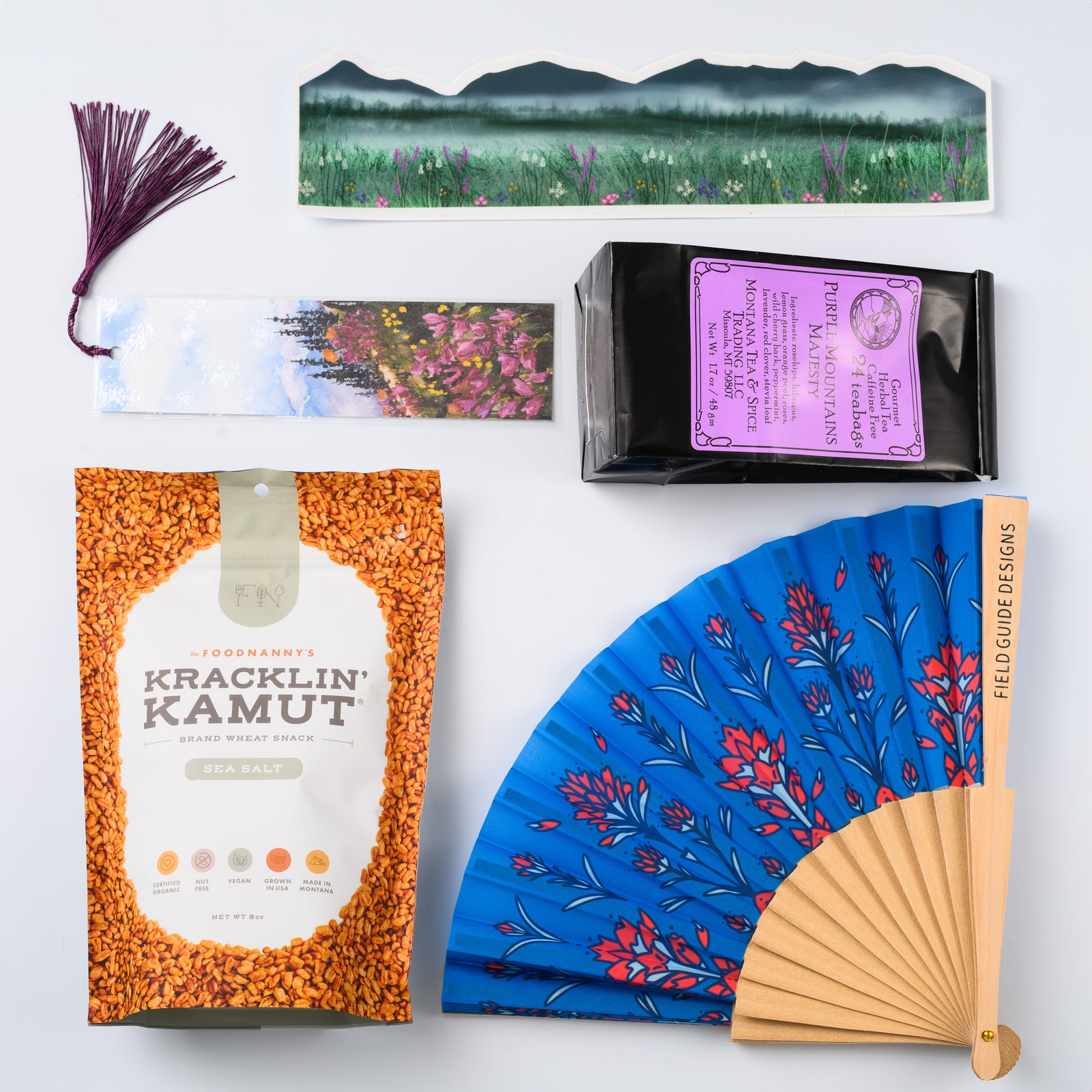 A flat lay image of a Montana made gift box featuring a bag of Kracklin Kamut khorasan wheat snacks, a prairie fire/ (Indian) paintbrush hand fan, a watercolor bookmark, a bag of Montana Tea & Spice Co.'s Purple Mountain Majesty tea, and a large wildflower sticker wrap.