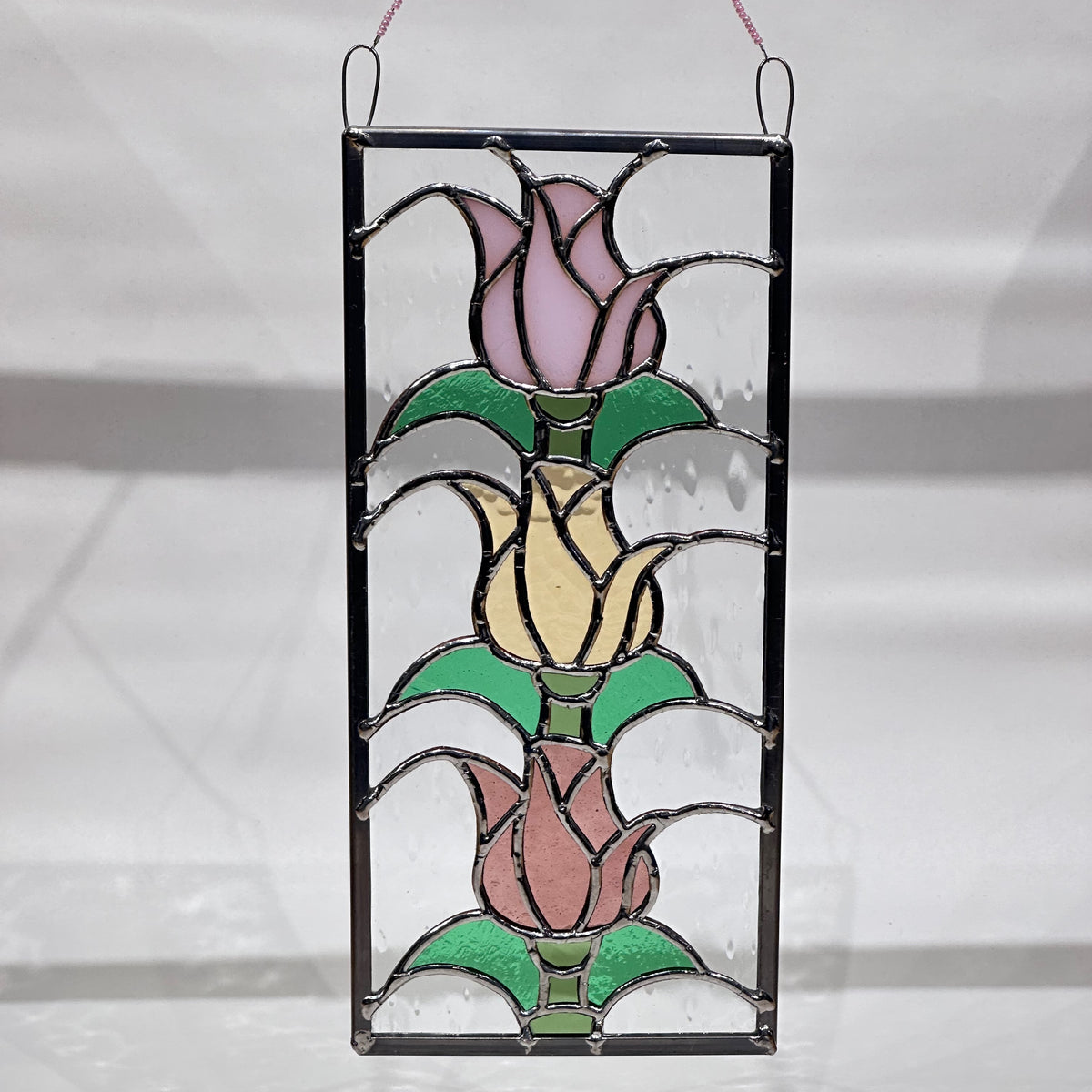 A stained glass piece with three stacked tulips in rose, yellow and pink glass in a rectangular frame.