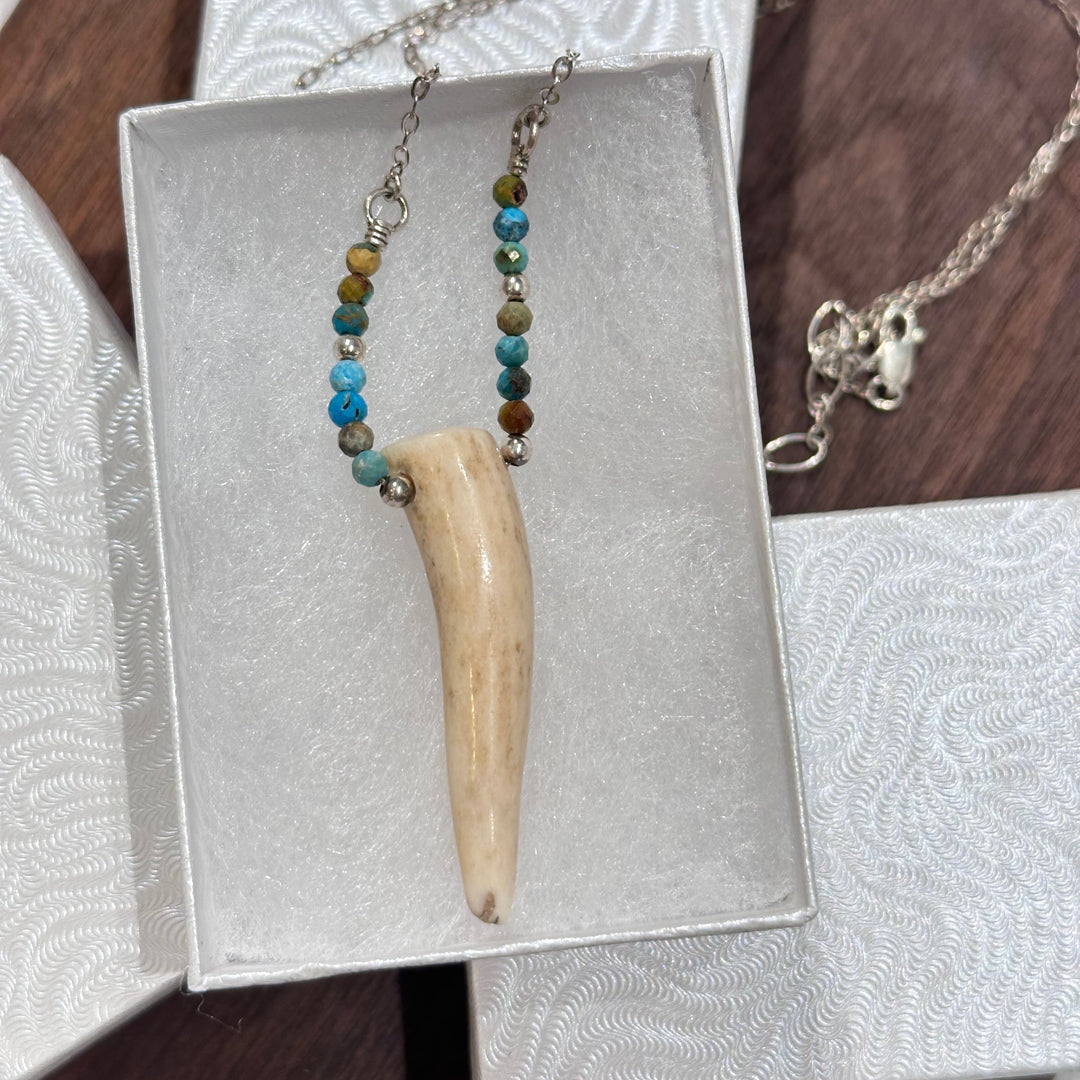 An antler tip necklace with turquoise and silver beads on a silver chain. Displayed in a white jewelry box on a a dark wood background.