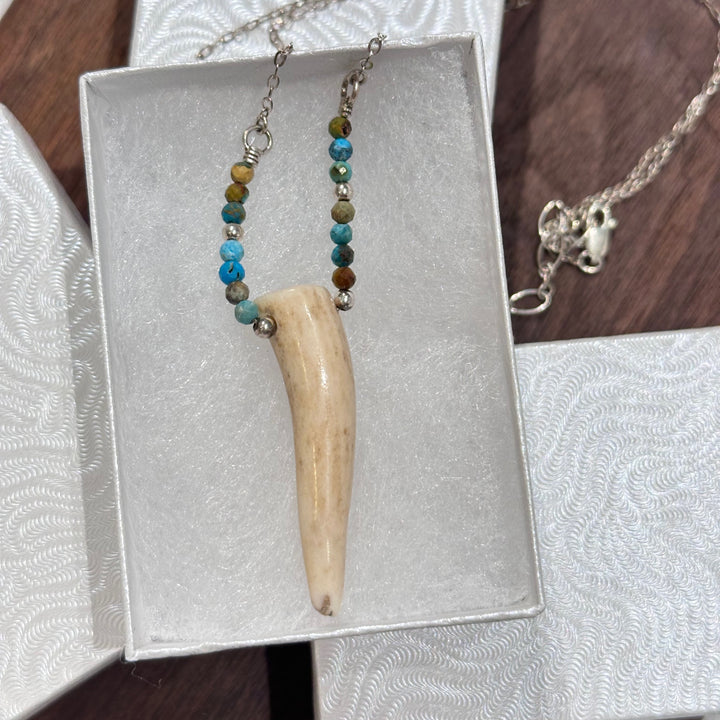 An antler tip necklace with turquoise and silver beads on a silver chain. Displayed in a white jewelry box on a a dark wood background.