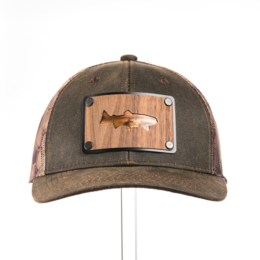 The front of a Kryptek trucker hat with a walnut wood trout cut patch out featuring bamboo patinated copper on a rectangular metal plate.
