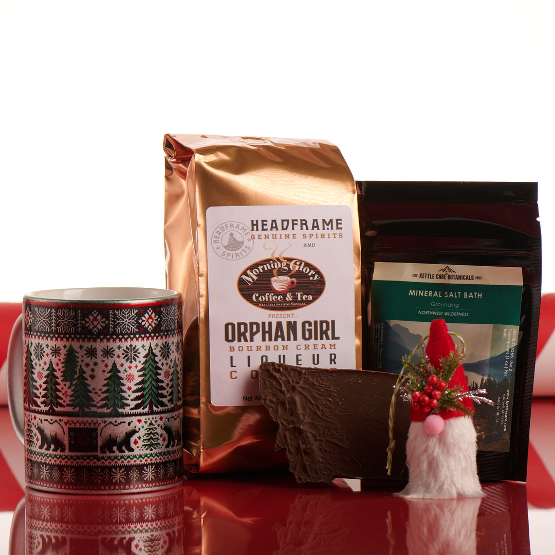 A picture of the Winter Wonderland box. A silver mug with a knit pattern of a bear, trees and snowflakes printed on it, a gnome ornament, a bag of orphan Girl Bourbon Cream Liqueur Coffee, a bag of Bath salt soak, and a milk chocolate bar in the shape of Montana