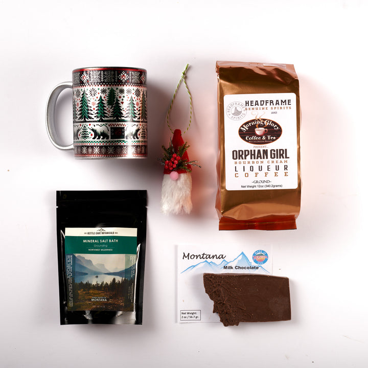 A flat lay of the Winter Wonderland box. A silver mug with a knit pattern of a bear, trees and snowflakes printed on it, a gnome ornament, a bag of orphan Girl Bourbon Cream Liqueur Coffee, a bag of Bath salt soak, and a milk chocolate bar in the shape of Montana
