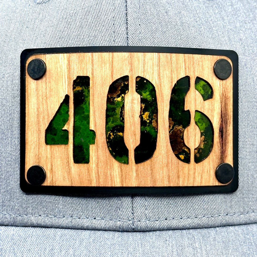 A close up of a Montana made wood patch riveted to a heather gray and black trucker hat with the 406 area code. Featuring green copper.