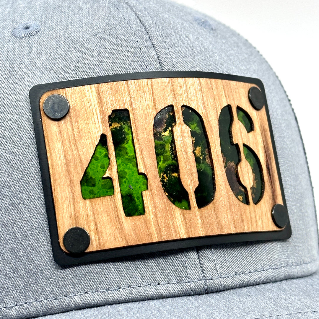 A Side view Montana made wood patch riveted to a heather gray and black trucker hat with the 406 area code. Featuring Green copper.