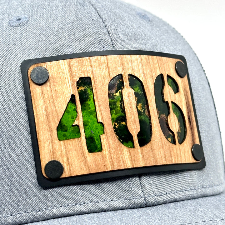 A Side view Montana made wood patch riveted to a heather gray and black trucker hat with the 406 area code. Featuring Green copper.
