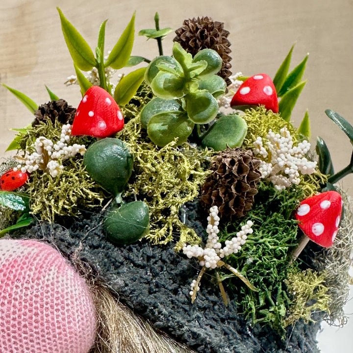 A gnome hat with a Decorative arrangement with moss, leaves, and red mushrooms on a textured surface.