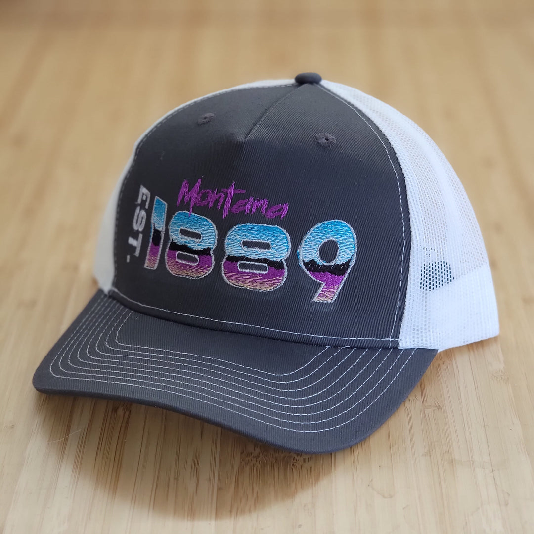  A picture of a steel grey trucker hat with white mesh and contrast stitching. It is embroidered with a design that reads Montana EST. 1889. The art style is retro 80s style.